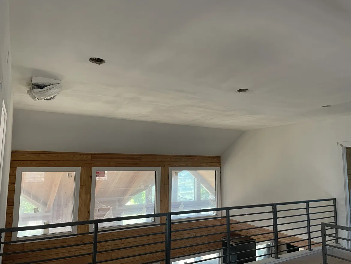 Upstairs loft with a sloped ceiling finished smooth and primed above a wood-paneled wall with three windows and a metal railing