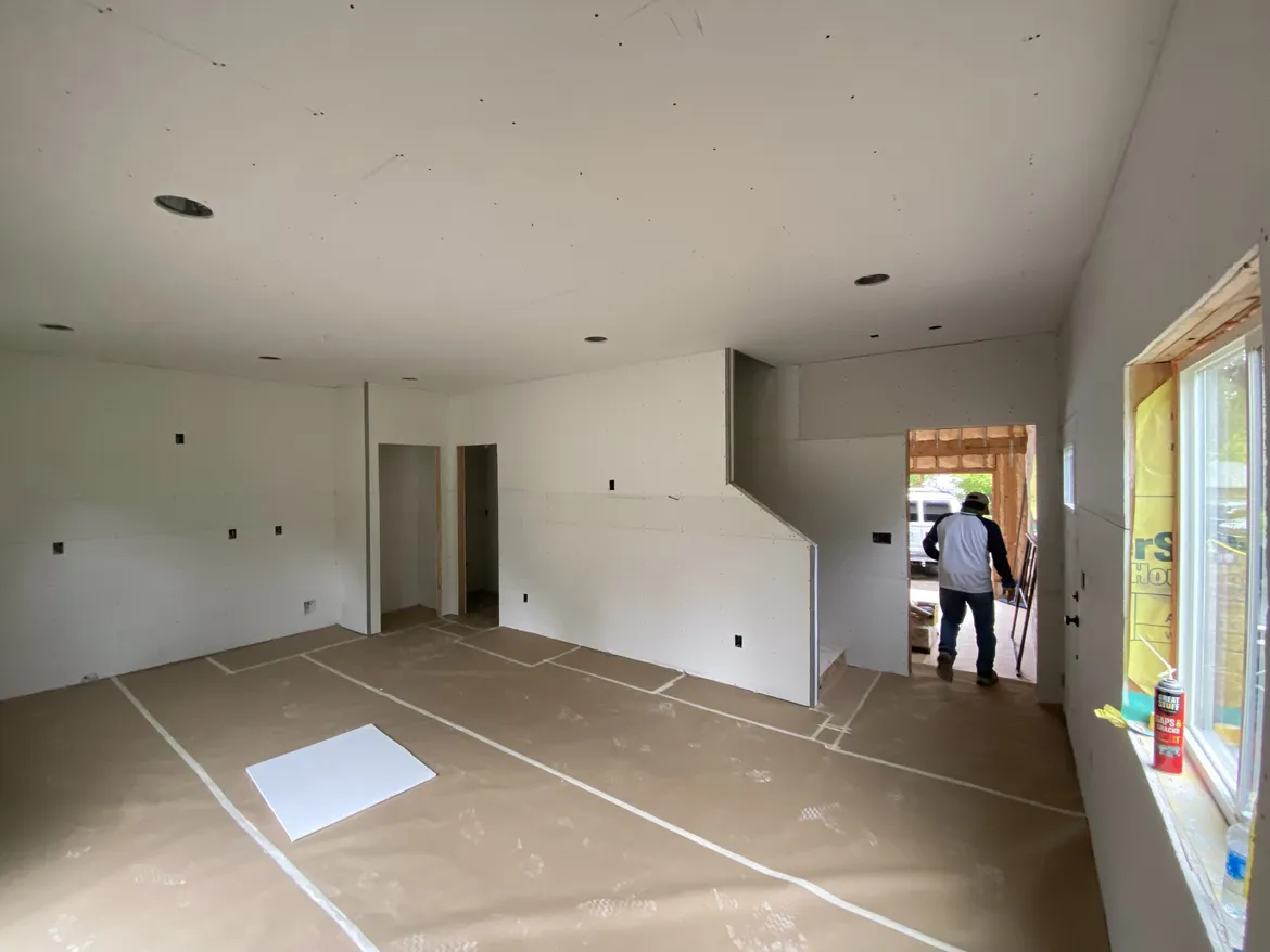 Room with drywall hung but not yet taped on the walls and ceiling, a stairway wall and closet openings, and a crew member walking out a doorway