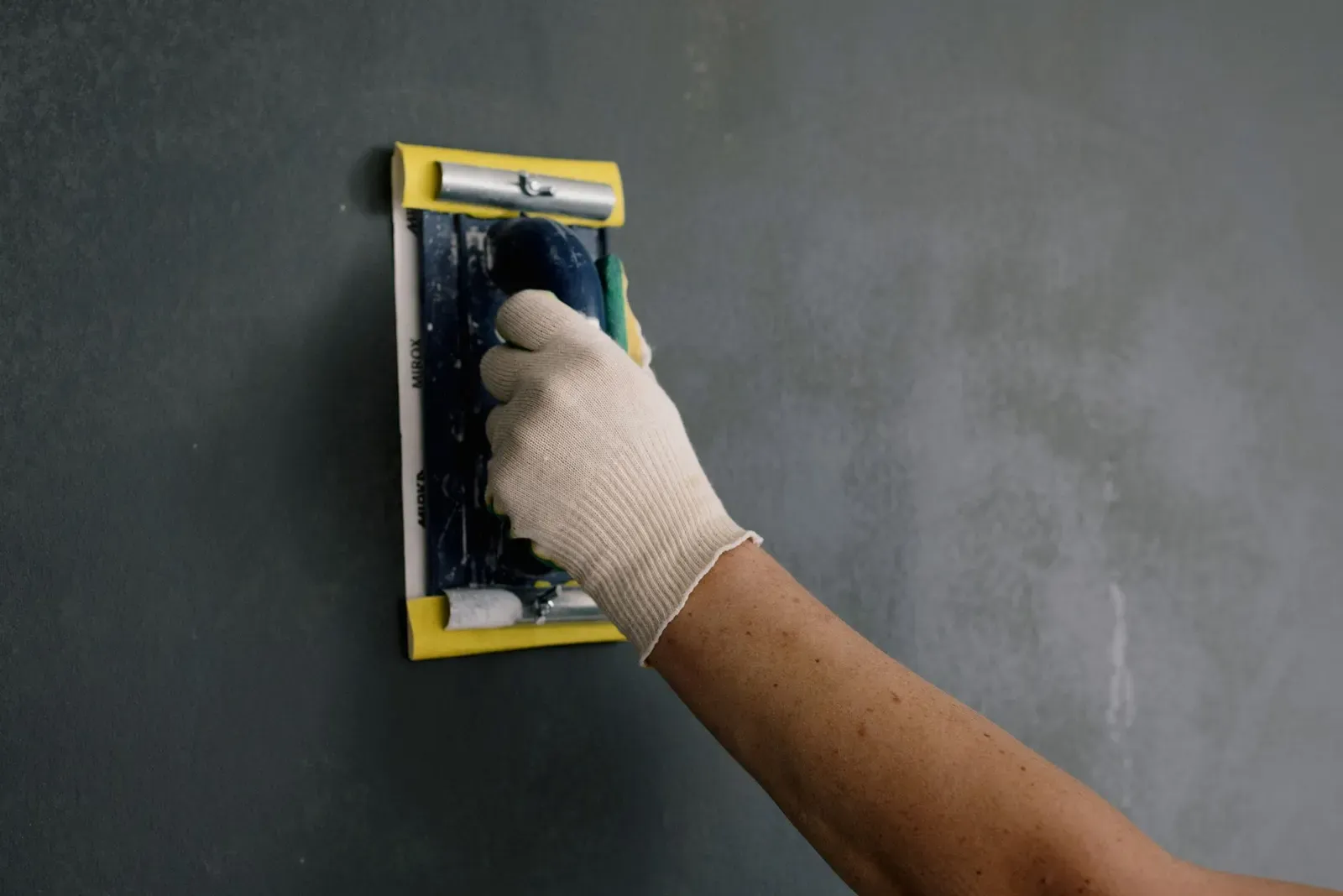 Gloved hand sanding a smooth wall with a hand sanding block after a drywall patch